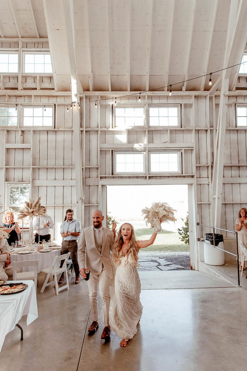 A couple enters their wedding reception in a bright white barn venue with windows and rustic decor.
