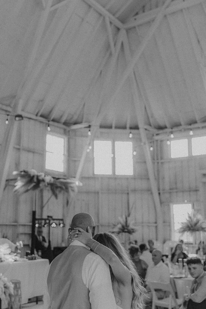 Black and white photo of a couple dancing at a wedding reception in a rustic venue with high ceilings and windows.