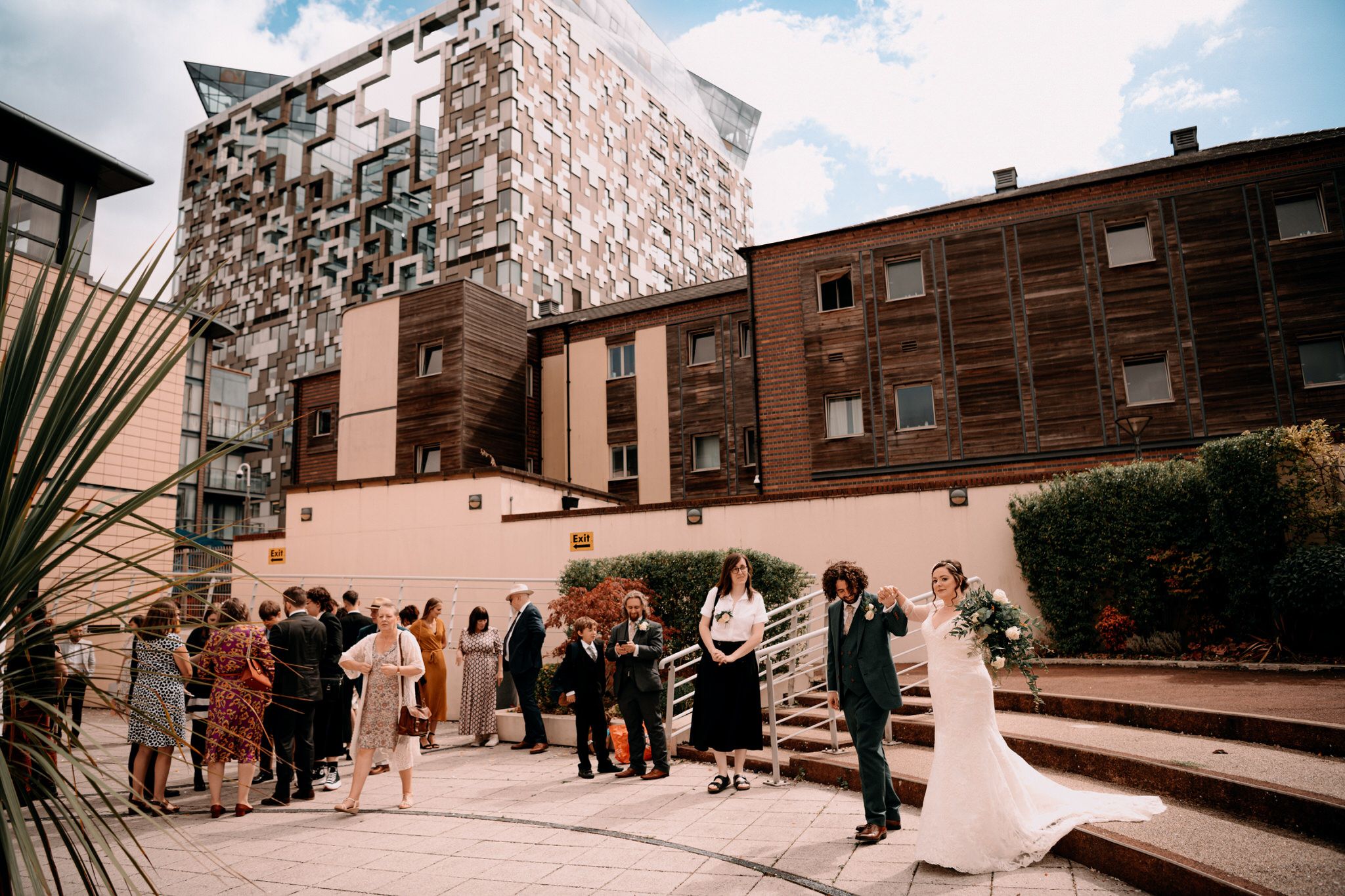 Wedding guests gather on an outdoor terrace with pink walls and modern architecture in the background.
