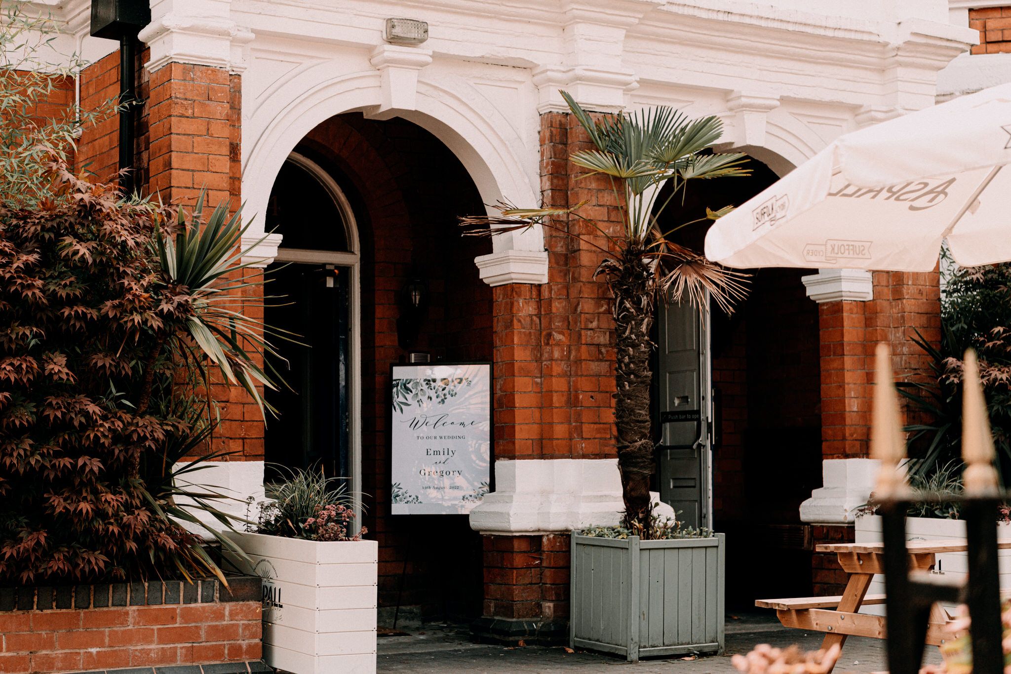 White arched entrance of a brick building with tropical plants and outdoor umbrella seating.