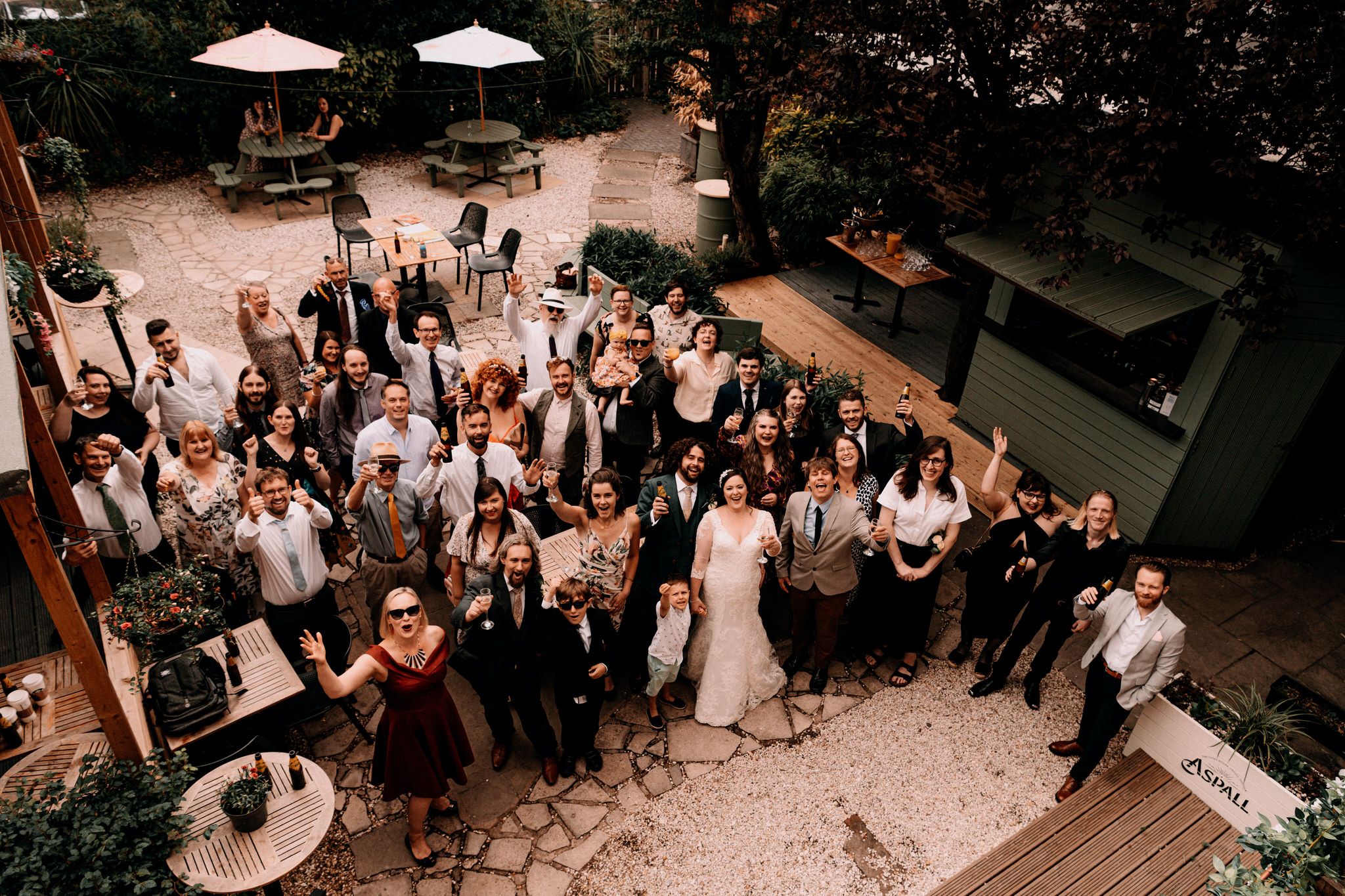 Aerial view of a wedding celebration on an outdoor patio with guests gathered around tables and umbrellas.