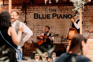 Musicians perform at The Blue Piano venue with acoustic guitar and upright bass against a rustic brick wall backdrop.