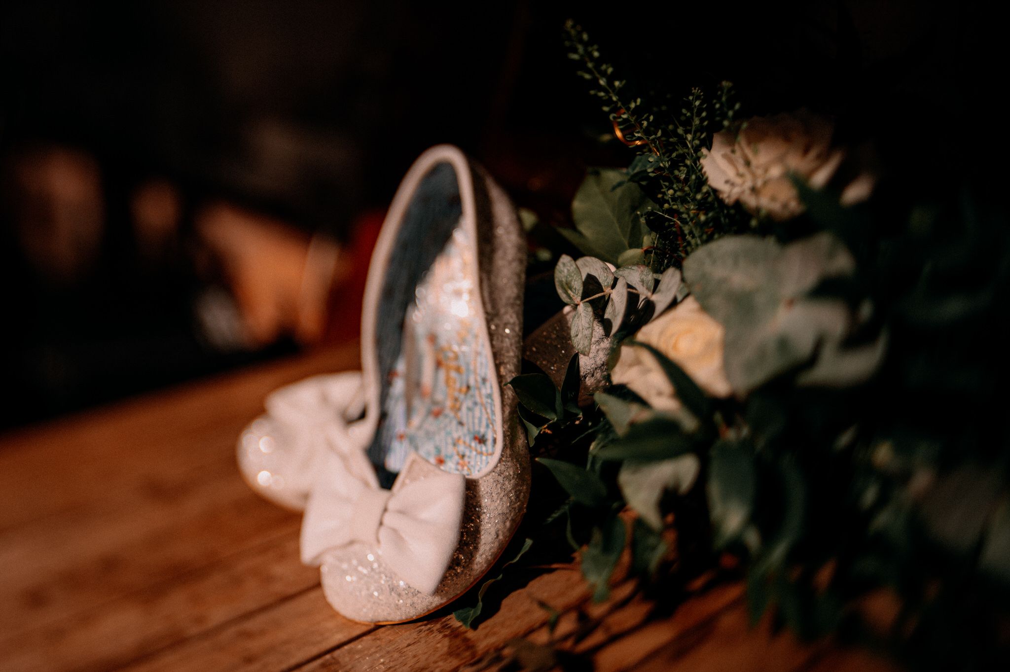 Beautiful lace wedding shoes on wooden surface next to floral arrangement in dark moody lighting.