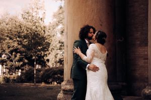 A romantic wedding couple embraces and kiss in an outdoor setting with stone pillars and lush greenery in the background.