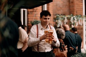 A guest in suspenders enjoys a beer at an outdoor wedding reception with brick walls in the background.