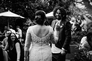 Black and white candid moment at an outdoor wedding reception with lace-backed dress and guests at tables and umbrellas.