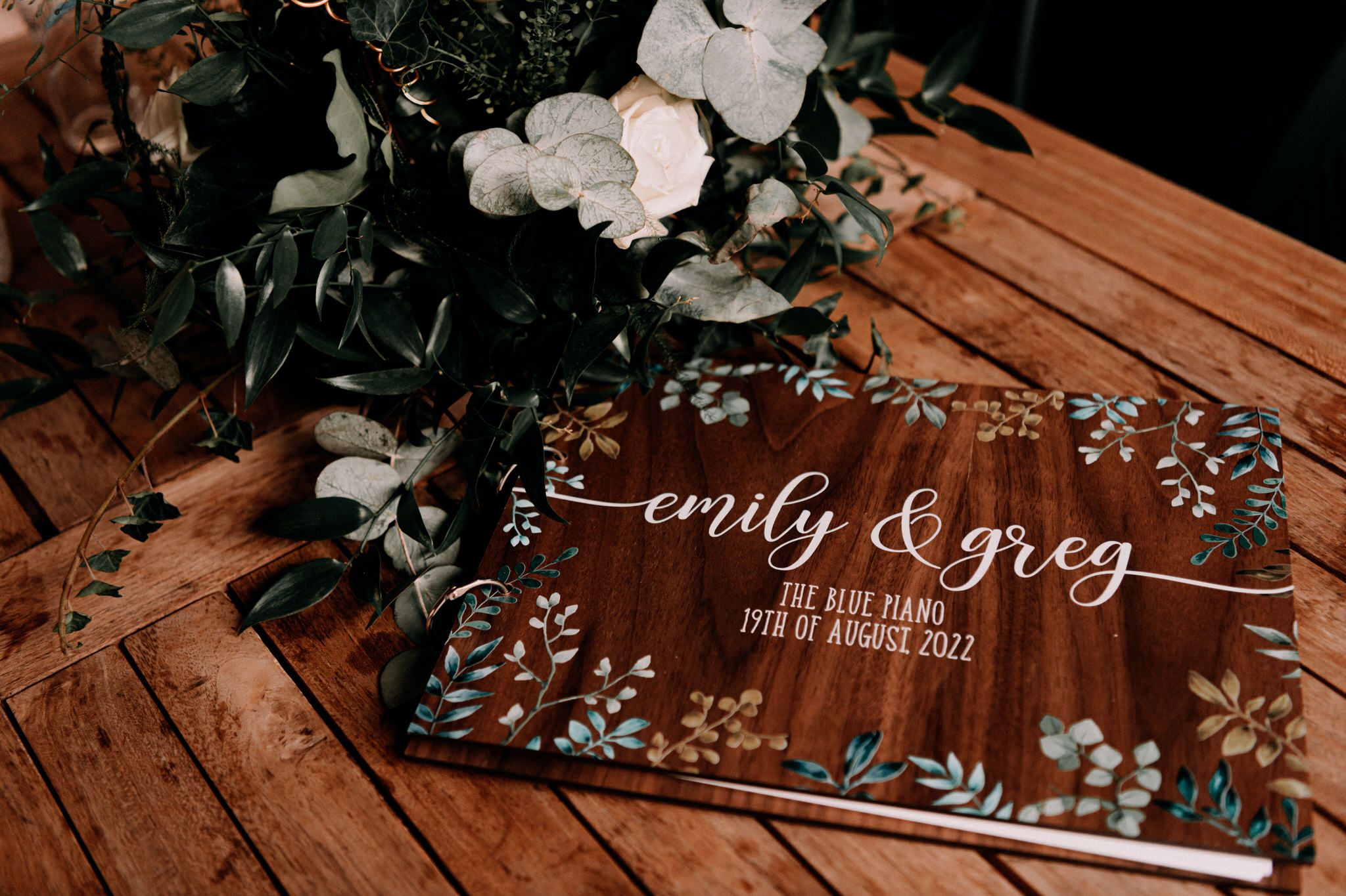Clear acrylic wedding invitation sign with white floral design and text on wooden surface beside white flowers.