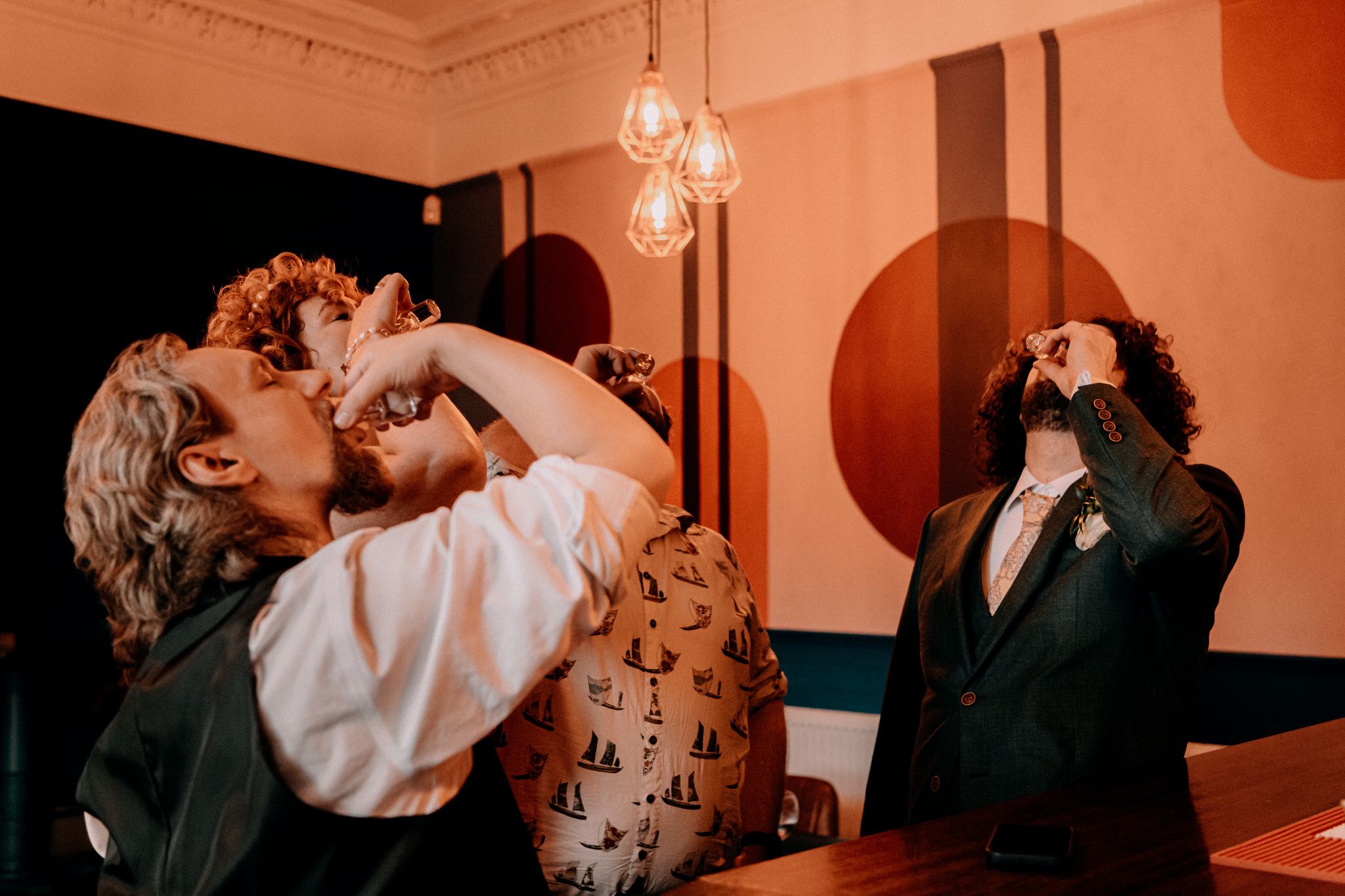 Two people drinking in sync at an ornate vintage bar with mood lighting and retro geometric wall design.