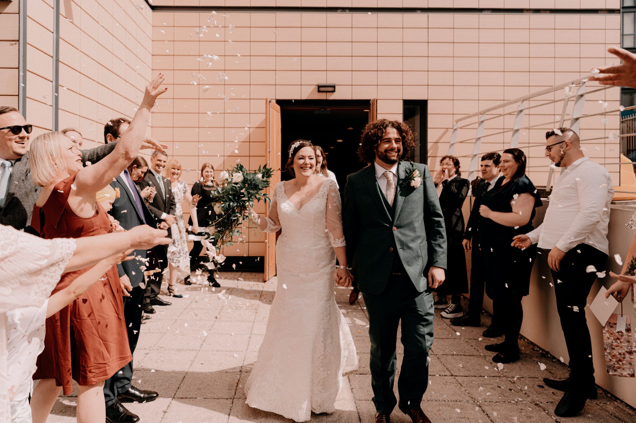Wedding couple exits ceremony venue while guests throw flower petals and celebrate along the aisle walkway.