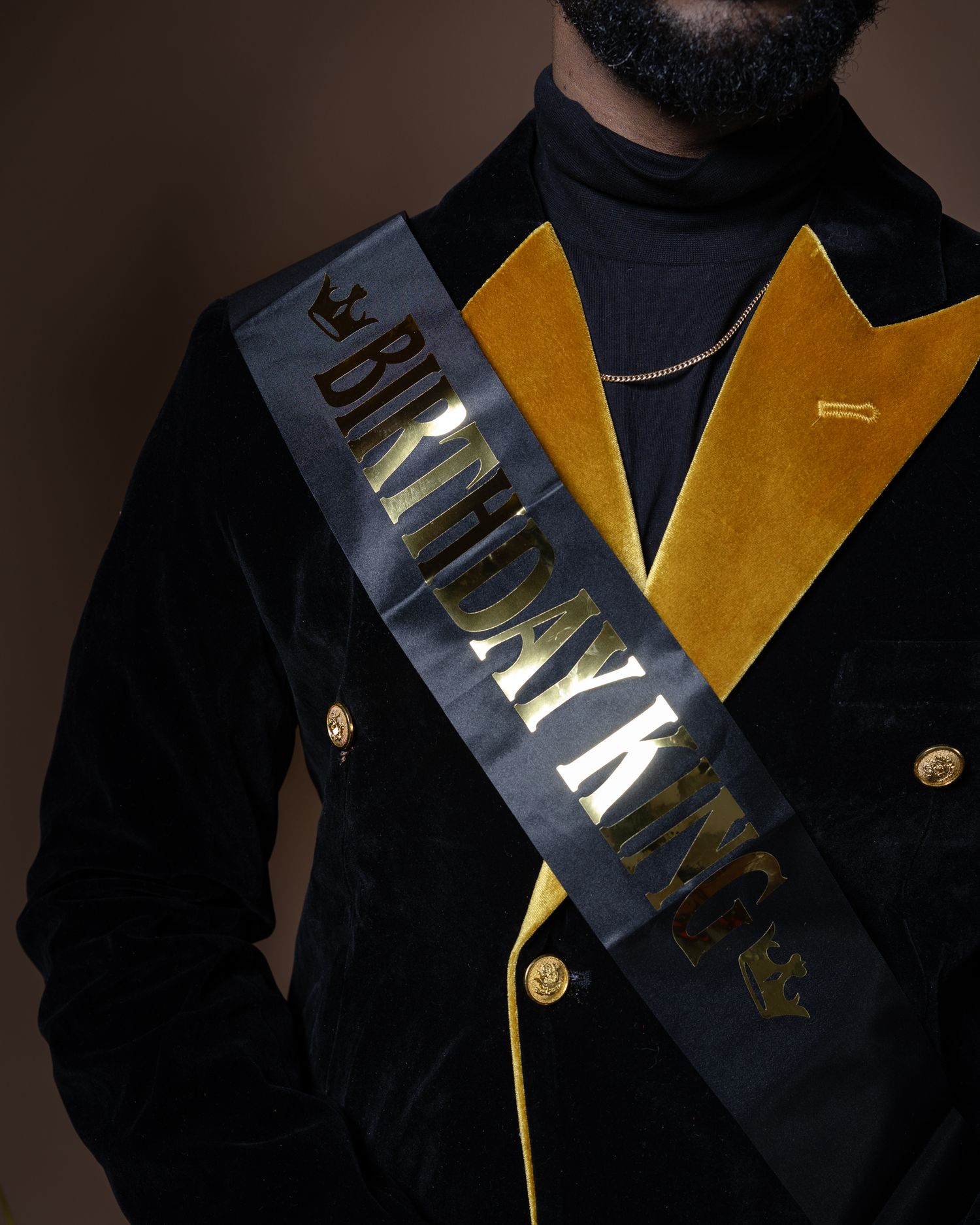 A black velvet blazer with gold lapels and buttons features a dark sash with gold lettering across the chest.