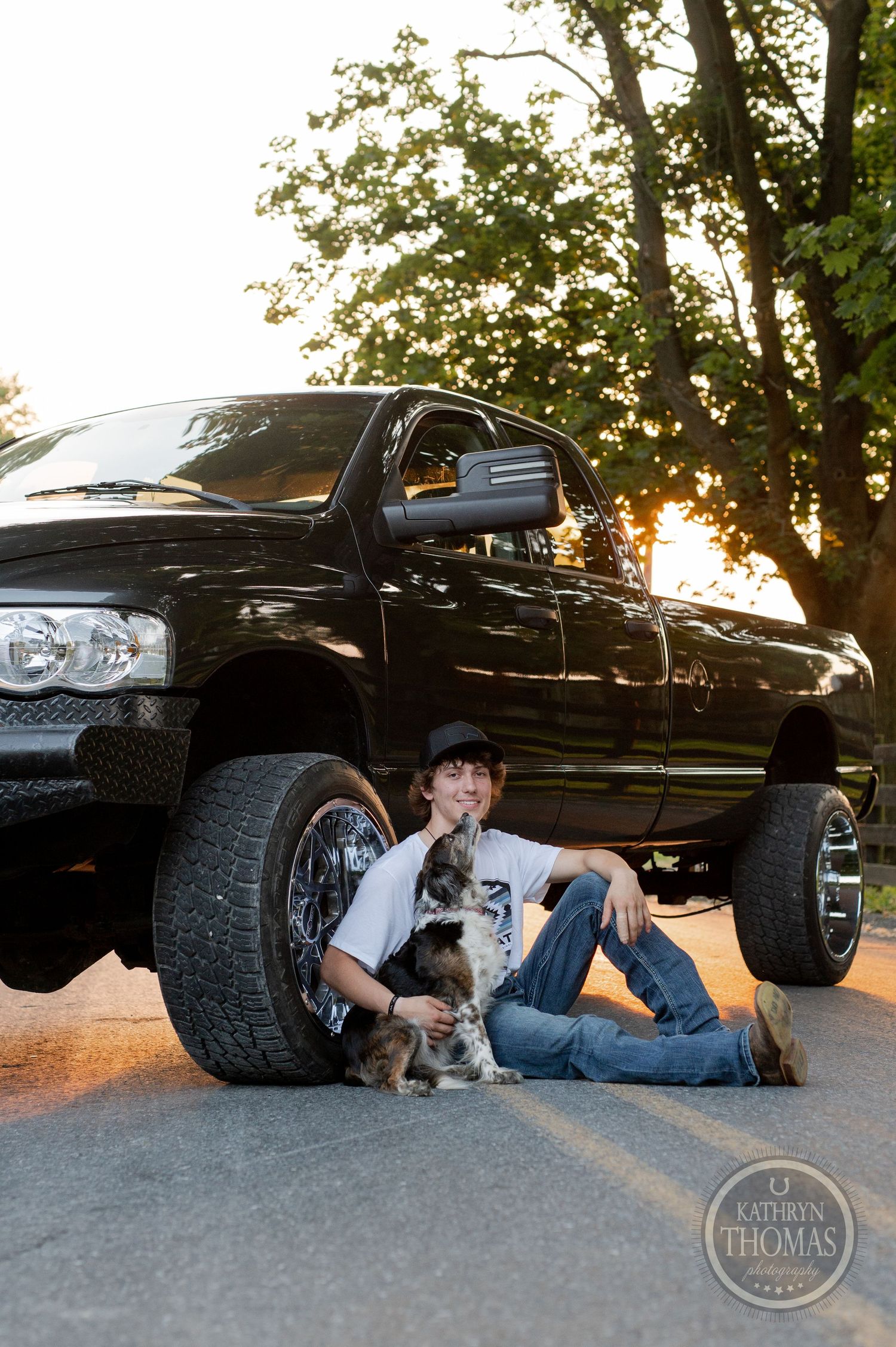 Lifted black Dodge Ram truck with a small dog against sunset backdrop on country road.