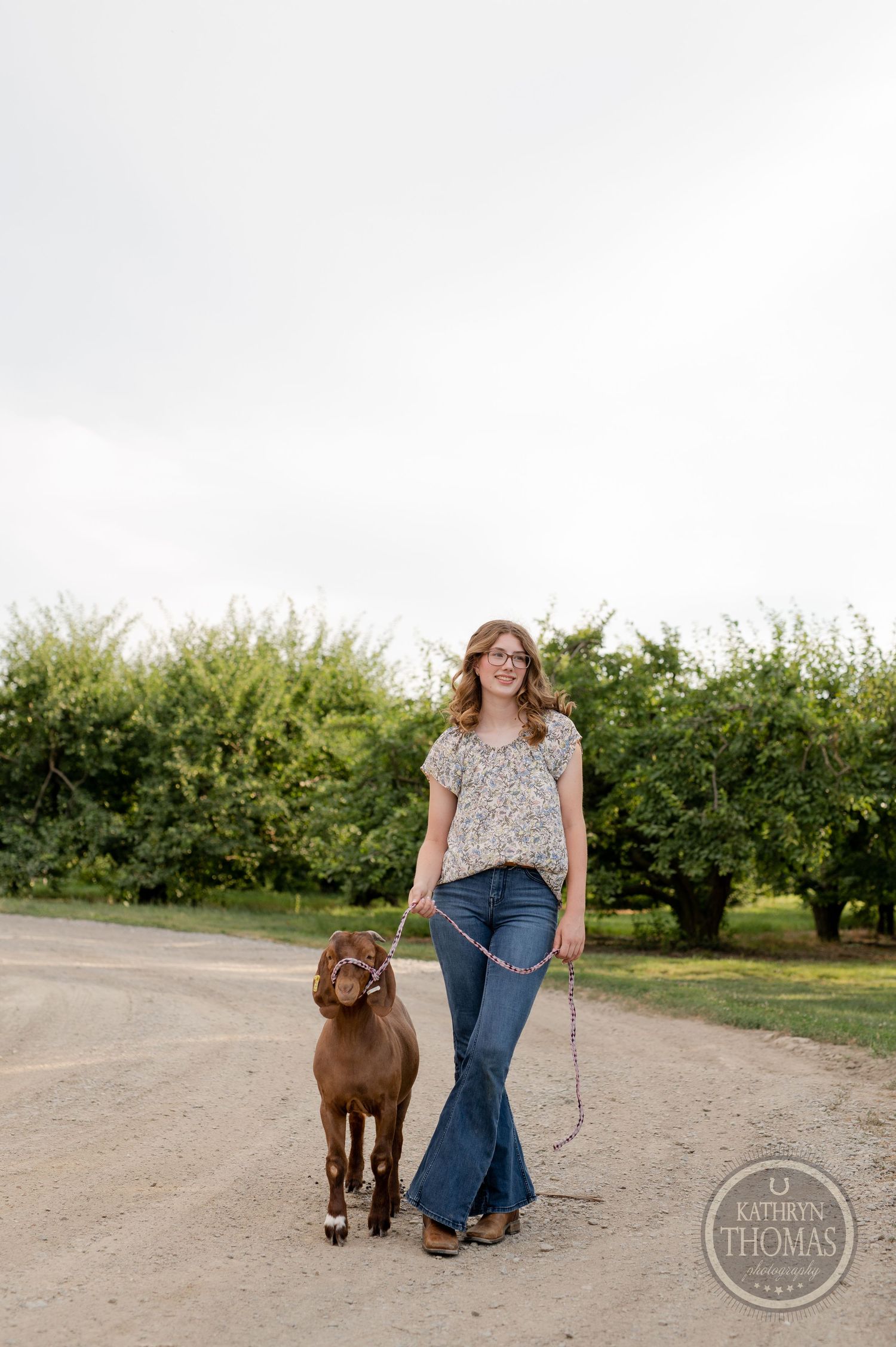Person and brown goat walking together on rural dirt path with greenery in background.