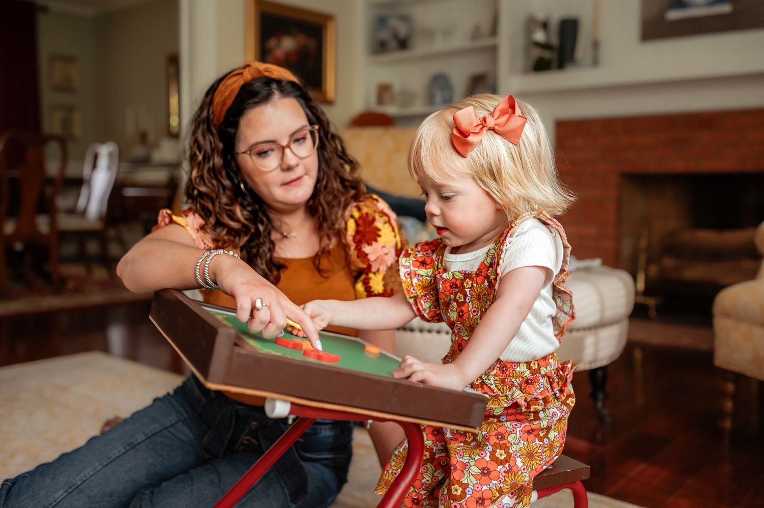 A small child and adult wearing floral dresses read a book together while sitting on the floor at home.
