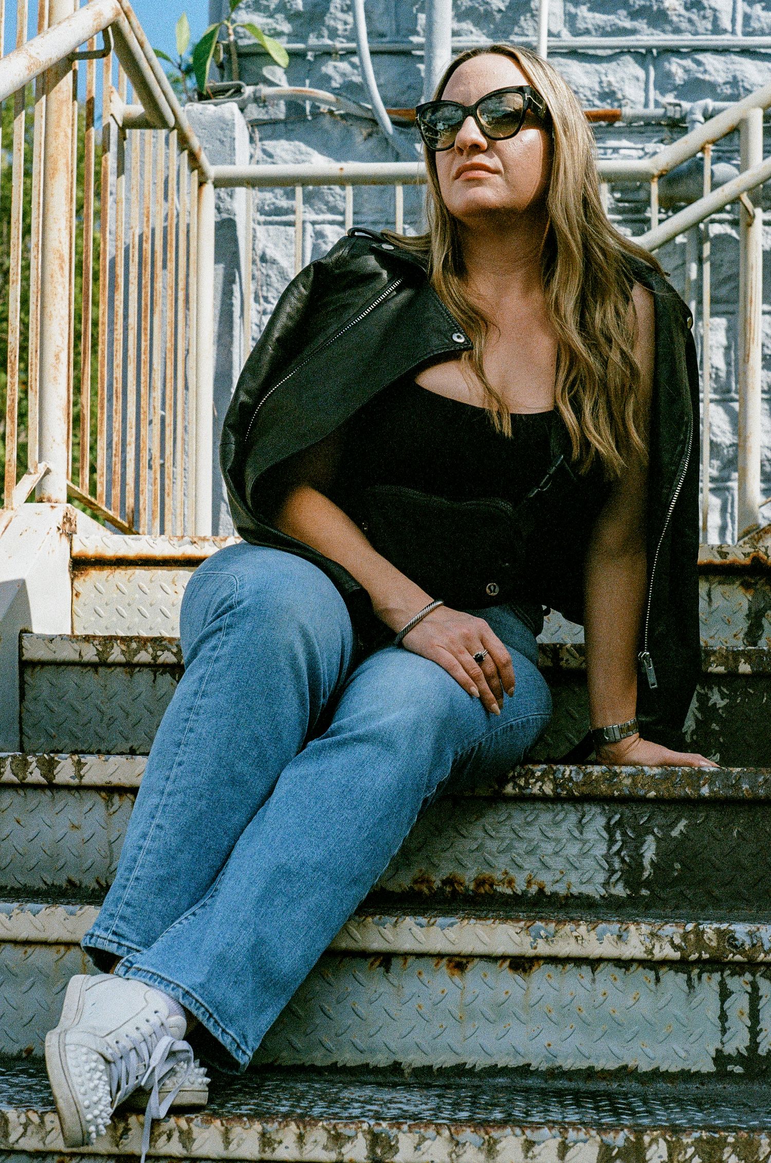 A casually dressed person in jeans, black jacket and sunglasses poses sitting on concrete steps outdoors.