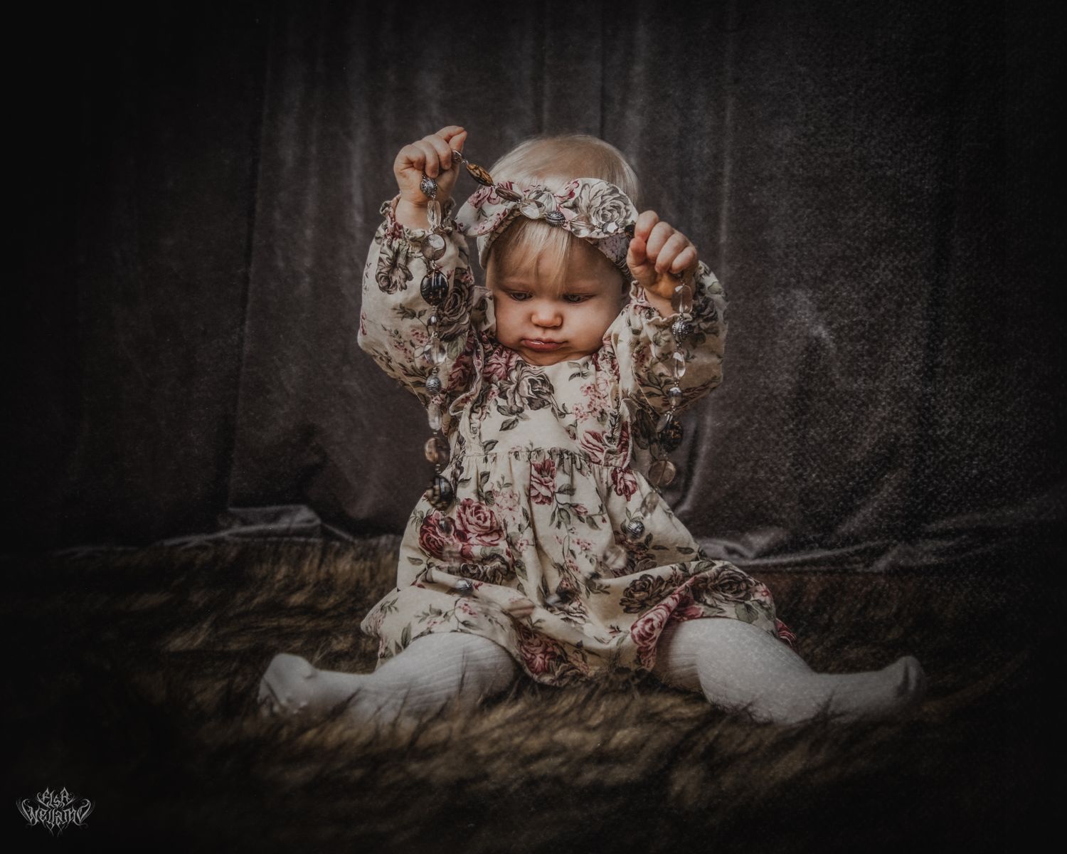Baby in floral outfit sits on textured blanket against dark backdrop with soft lighting.