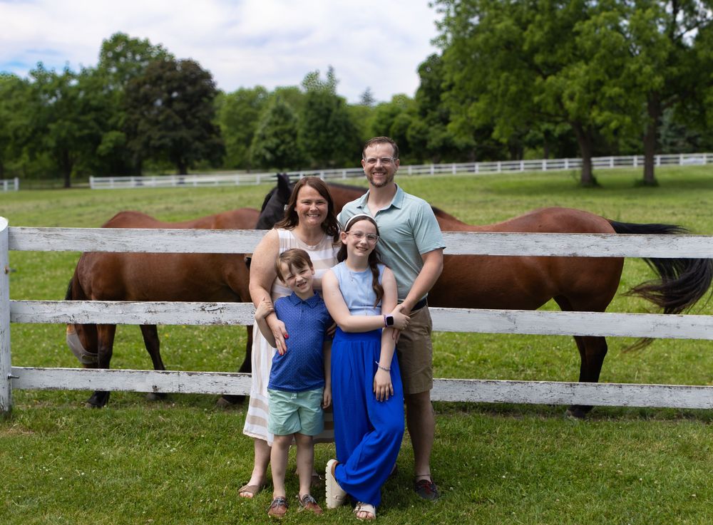 Family of four smiling in front of white fence with brown horses on green farm.