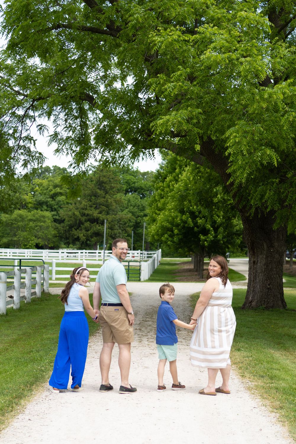 Family of four holding hands on a scenic gravel path surrounded by lush green trees and white fences.
