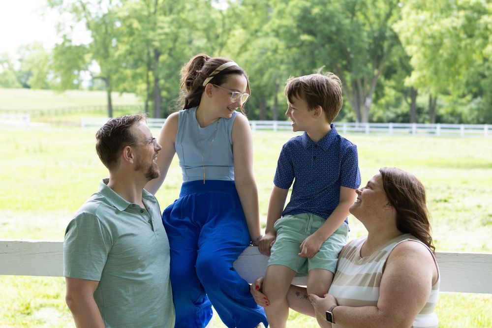 A happy family of four laughing together outdoors on a sunny day near a white fence surrounded by green trees.