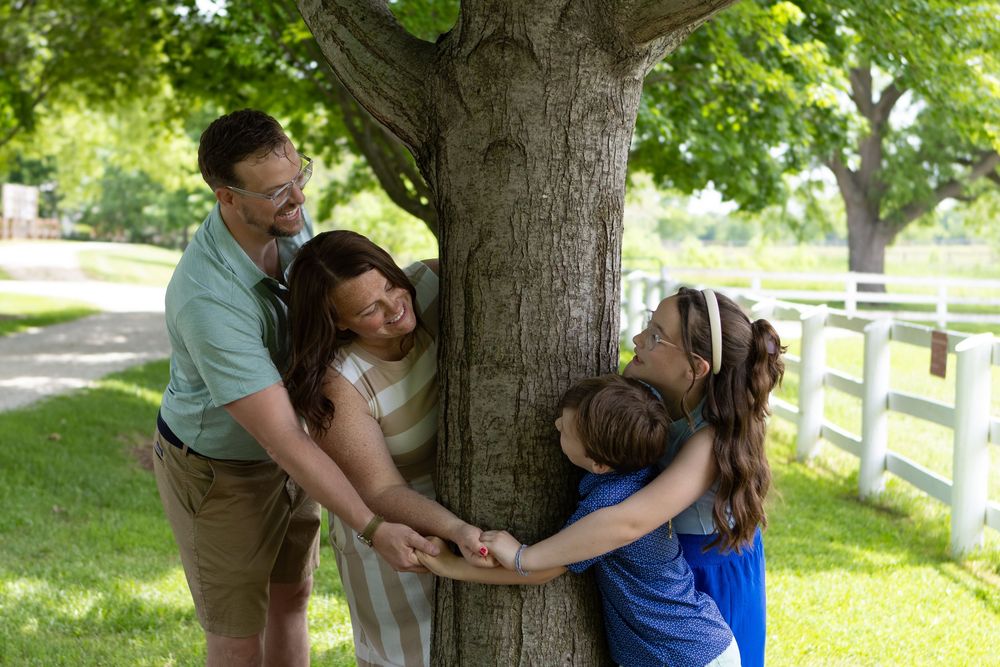 Joyful family hugging a large tree trunk in a sunny green park with white fences.