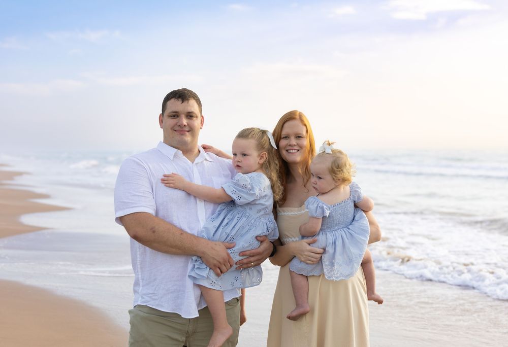 Family of four smiling on a sandy beach, parents holding two toddlers in matching blue dresses at sunset.