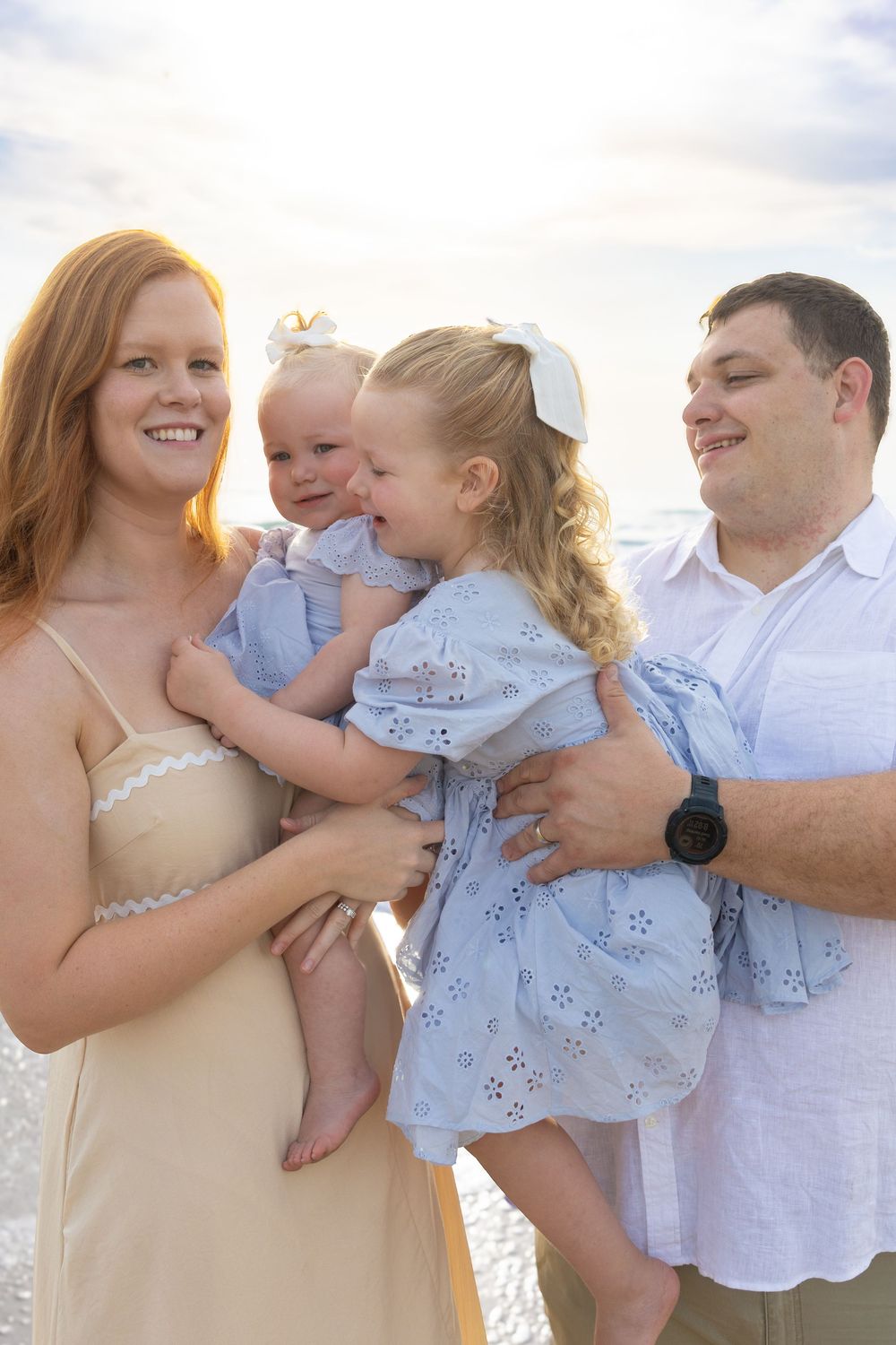 Happy family of four posing on a beach at sunset, parents holding two young daughters in matching blue dresses.