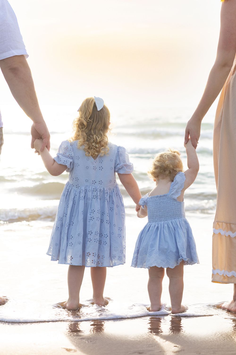 Two young girls in matching blue dresses hold hands with parents on a sunny beach shore.