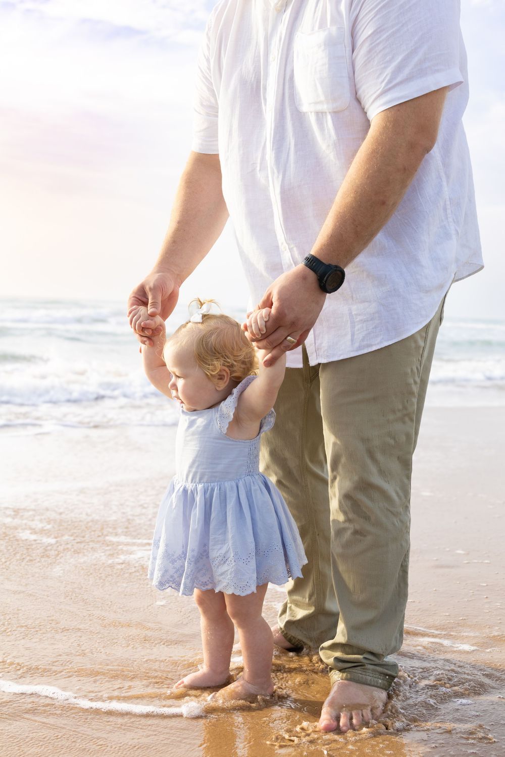 Father helps toddler girl walk along sandy beach shoreline, waves washing over their bare feet at sunset.
