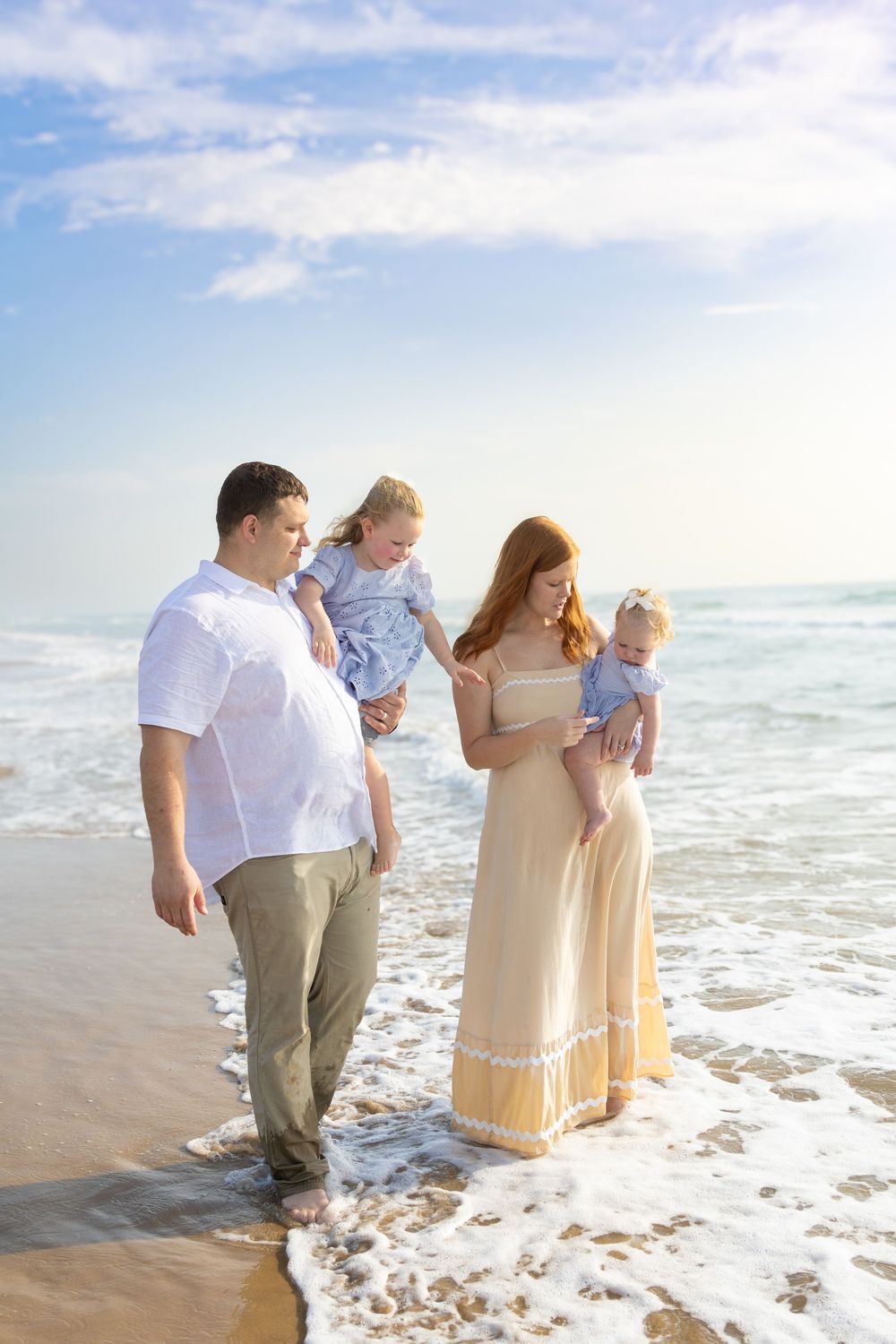 Happy family of four enjoying a beach walk, parents carrying young children along the ocean shoreline.