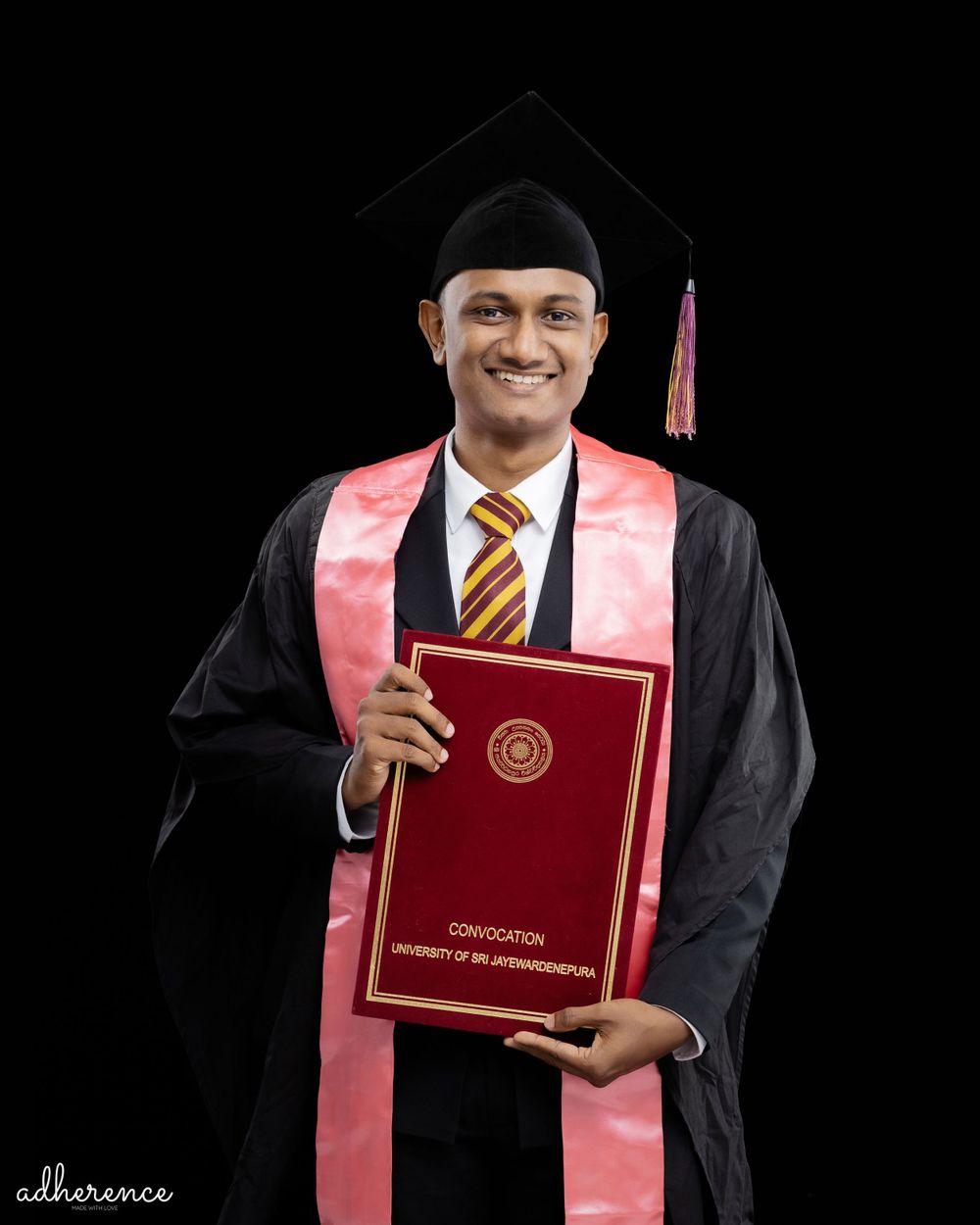 Graduate in black and pink academic regalia proudly displays red diploma against dark background.