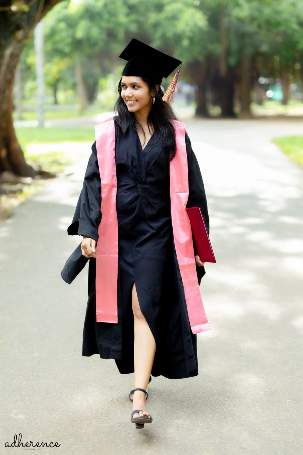 Graduate walks confidently in black academic regalia with pink stole on tree-lined path.