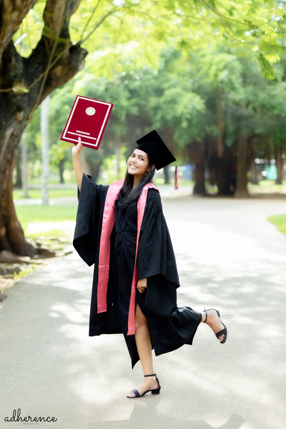 Graduate in black cap and gown playfully poses with diploma on campus pathway.