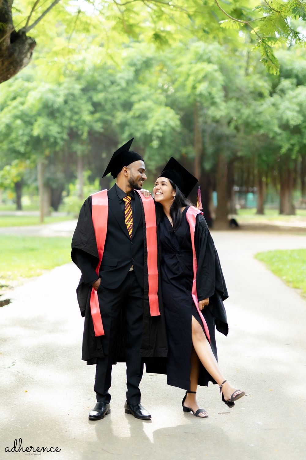 Two graduates in academic regalia share a candid moment on tree-lined campus walkway.