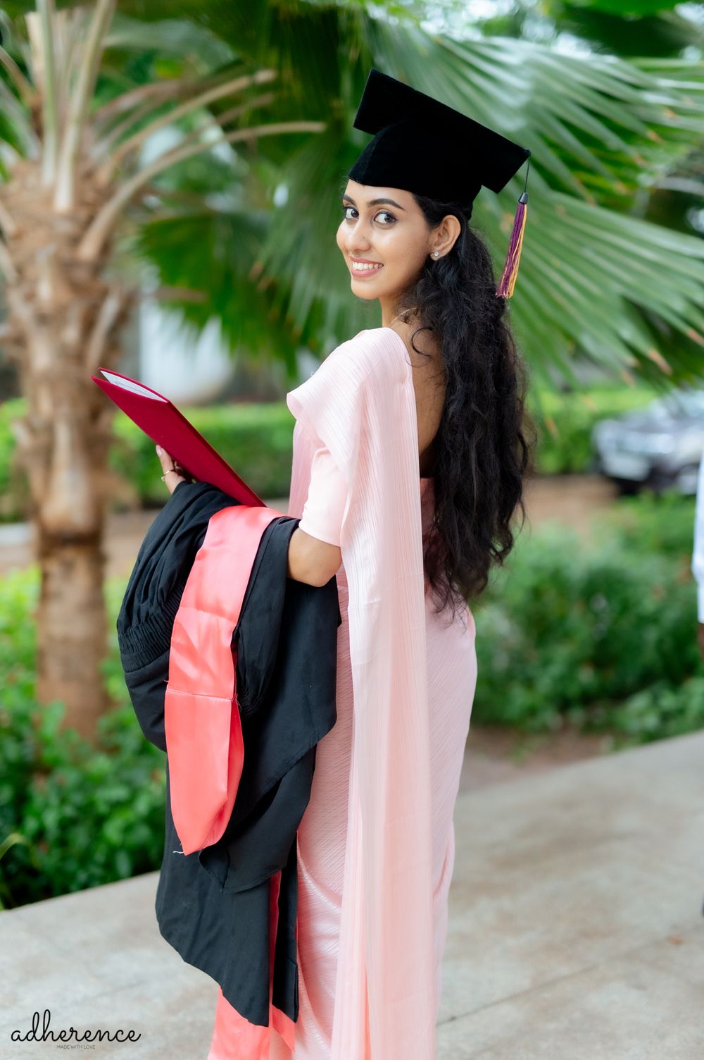 Graduate in black cap and gown with pink stole poses outdoors near palm trees.
