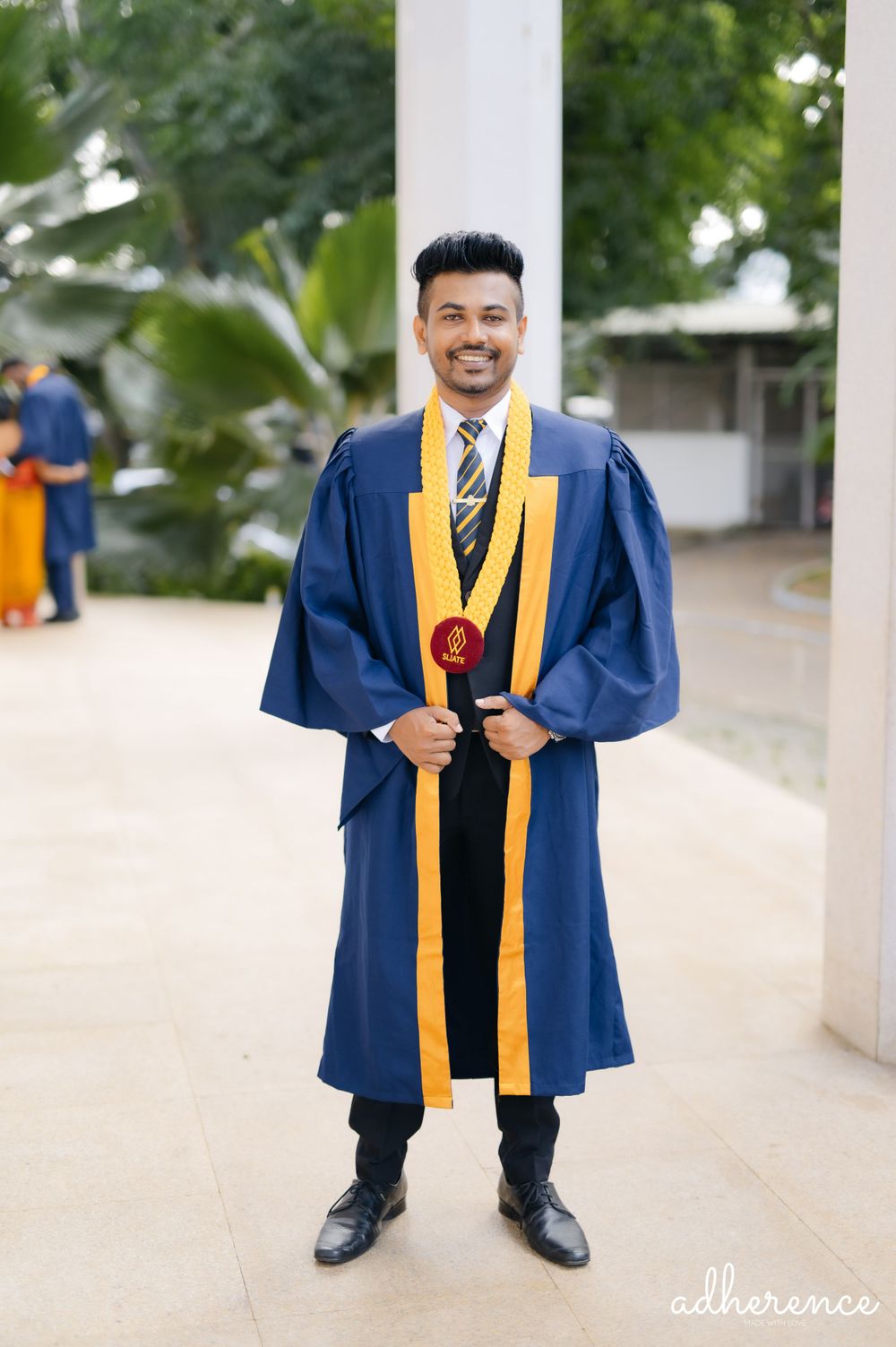 Graduate in blue and gold academic regalia stands proudly outdoors during commencement.