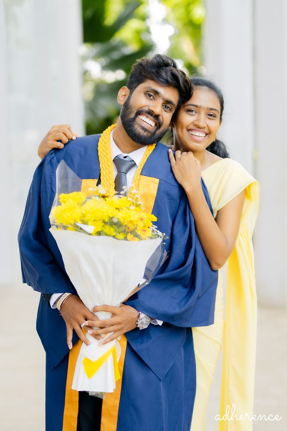 Graduate in blue academic regalia holding yellow flower bouquet poses with companion in yellow dress.