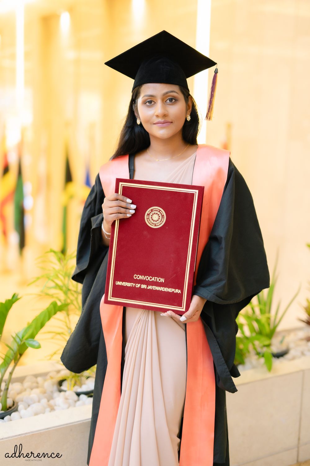 Graduate in black cap and gown holds up red diploma while standing against golden background.