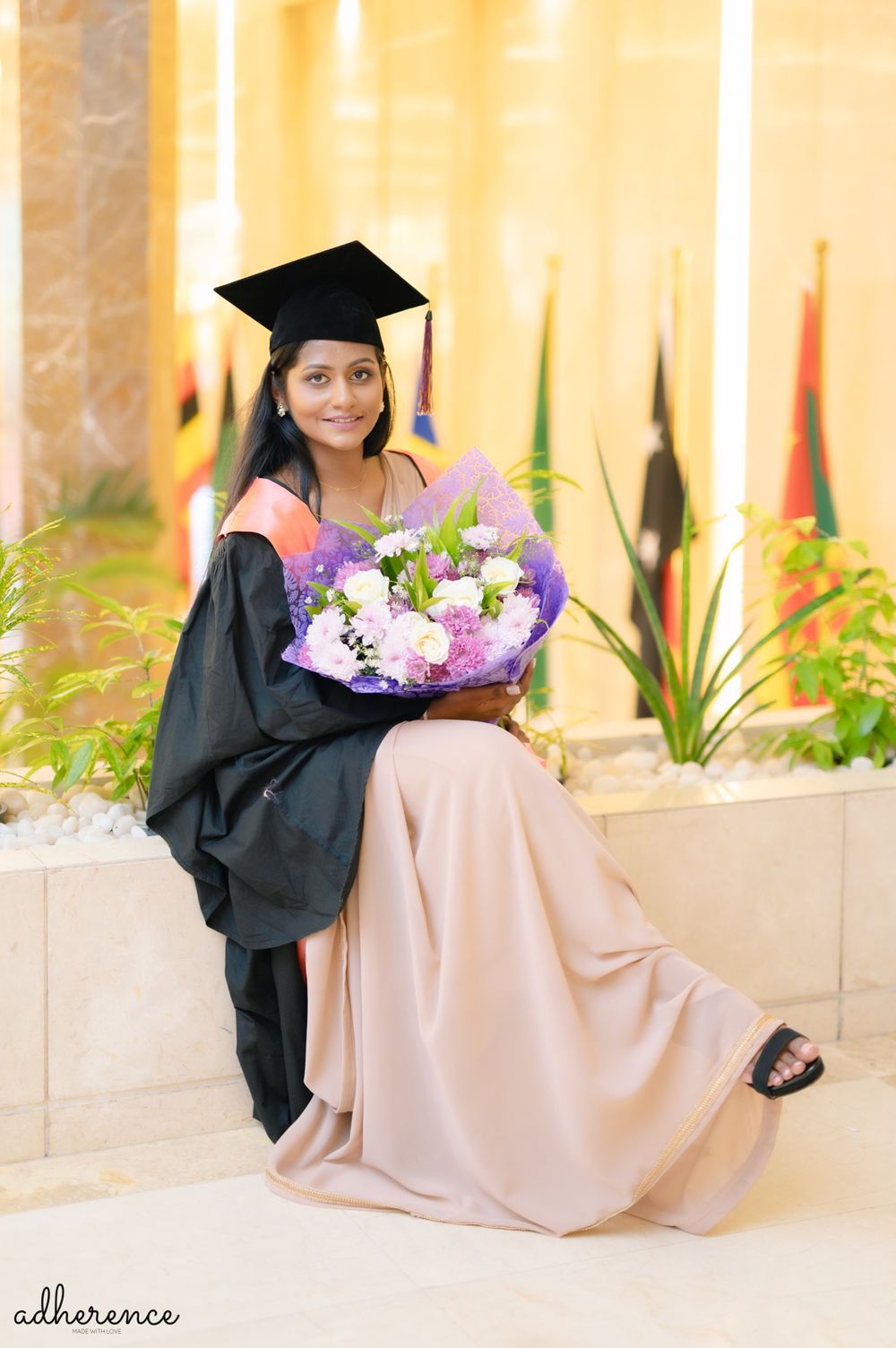 Graduate in black academic regalia poses with purple flower bouquet against warm-toned background.