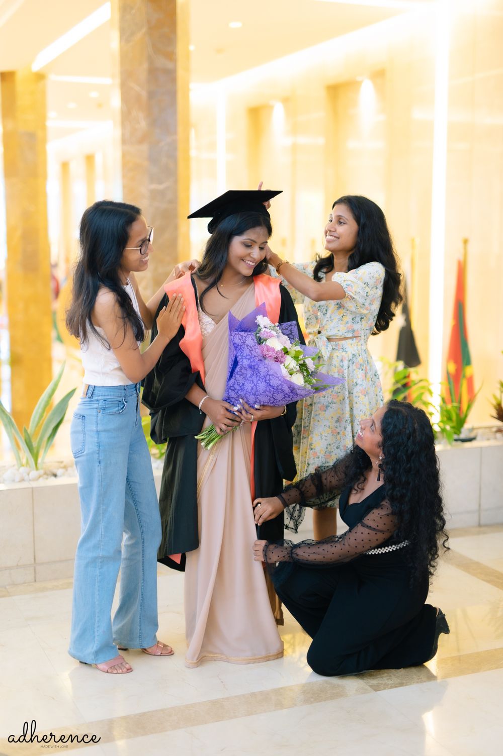 Friends gather around graduate with flowers for celebratory photos in warm-lit hallway.