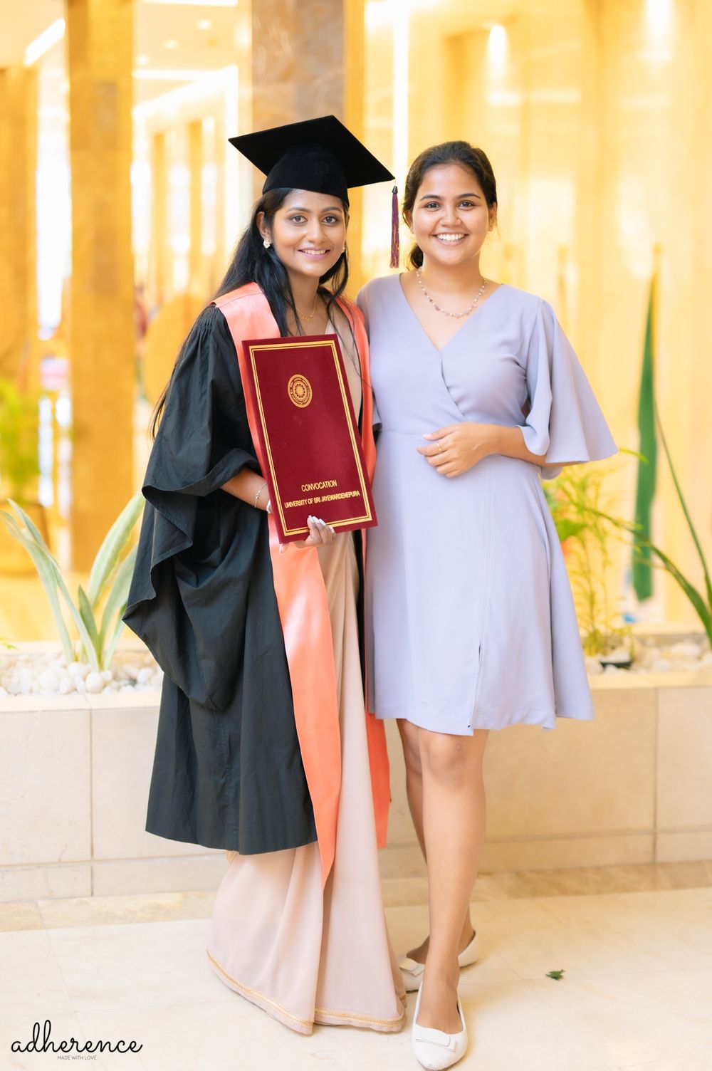 Two graduates pose together during commencement, one wearing traditional black cap and gown holding a diploma.