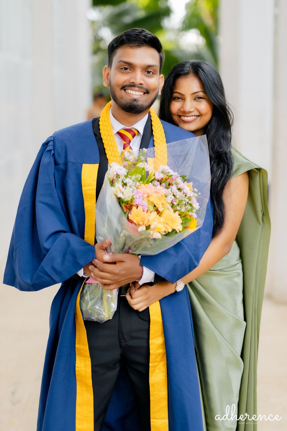 Graduate in blue and gold academic regalia holding colorful bouquet poses with companion in traditional dress.