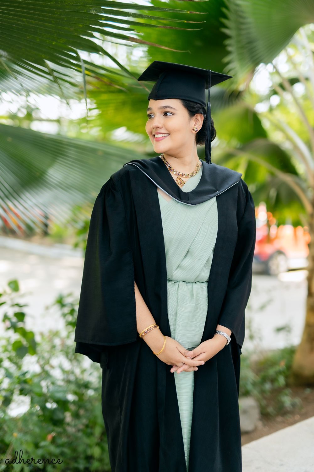 Graduate in black cap and gown with teal dress smiles outdoors among tropical palm fronds during graduation photo session.