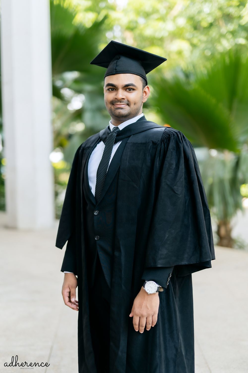 Graduate in traditional black academic regalia with cap and gown poses outdoors against green foliage.
