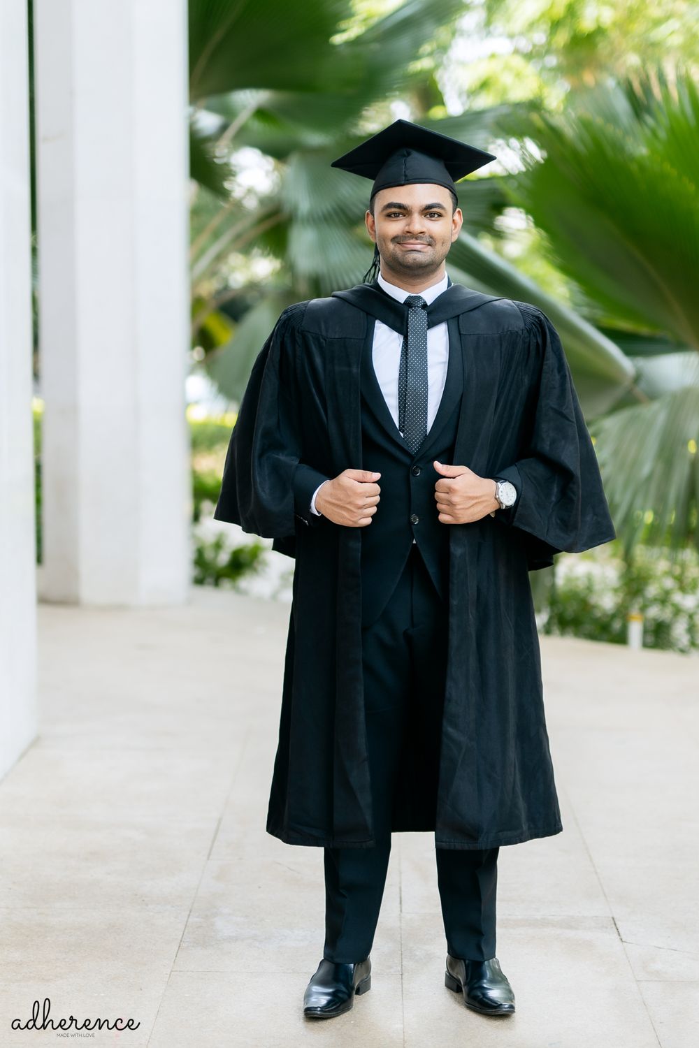 A graduate wearing traditional black academic regalia with cap and gown poses outdoors near tropical foliage.