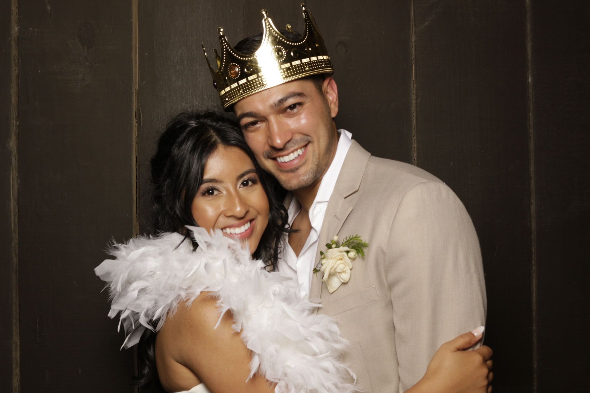 Couple poses happily at a wedding photo booth, man wearing a gold crown and woman with a white feather boa.