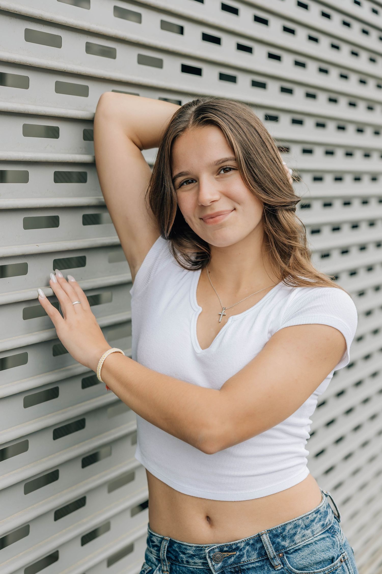 Young teenage girl in white crop top and jeans leaning against a gray slatted wall smiling at camera.