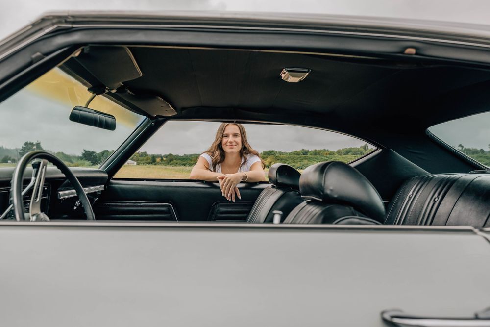 Woman leans over the front seat of a classic convertible car, smiling outdoors on a cloudy day.