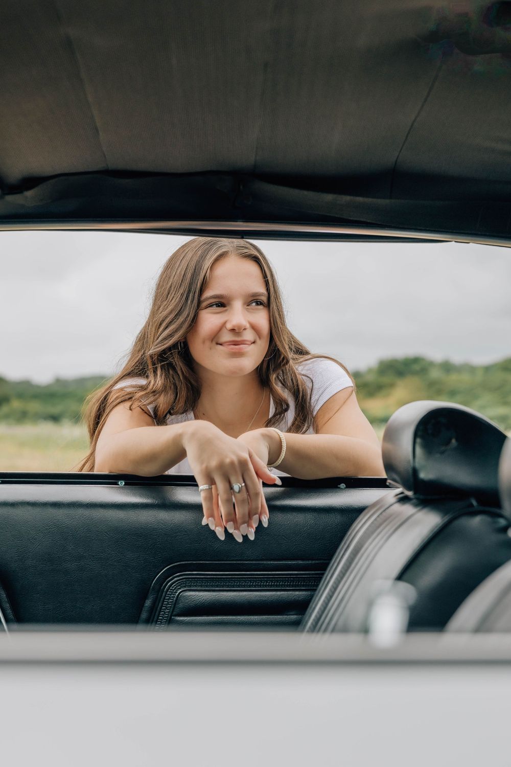 Young woman with long brown hair leans over car door of classic convertible, smiling softly indoors.