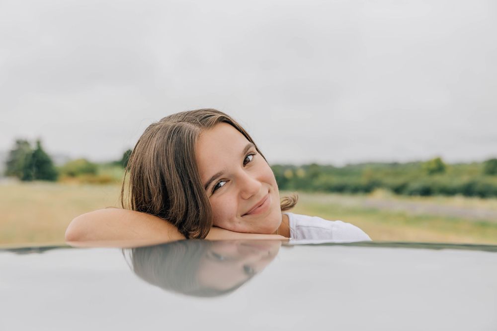 Young woman rests her head on the roof of a silver car, smiling gently with a field in background.