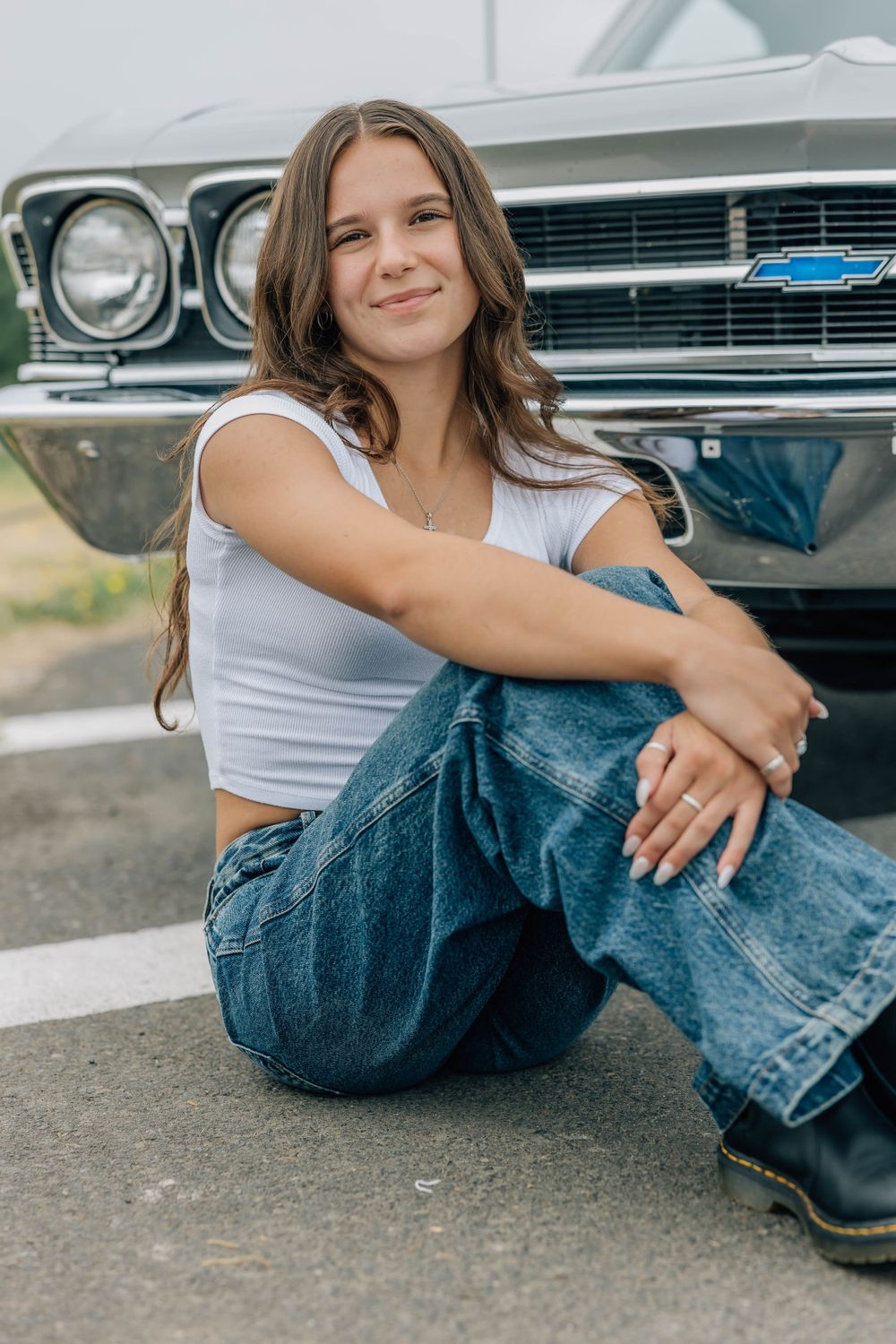 Teen girl in white crop top and jeans sits in front of a classic Chevrolet car on pavement.