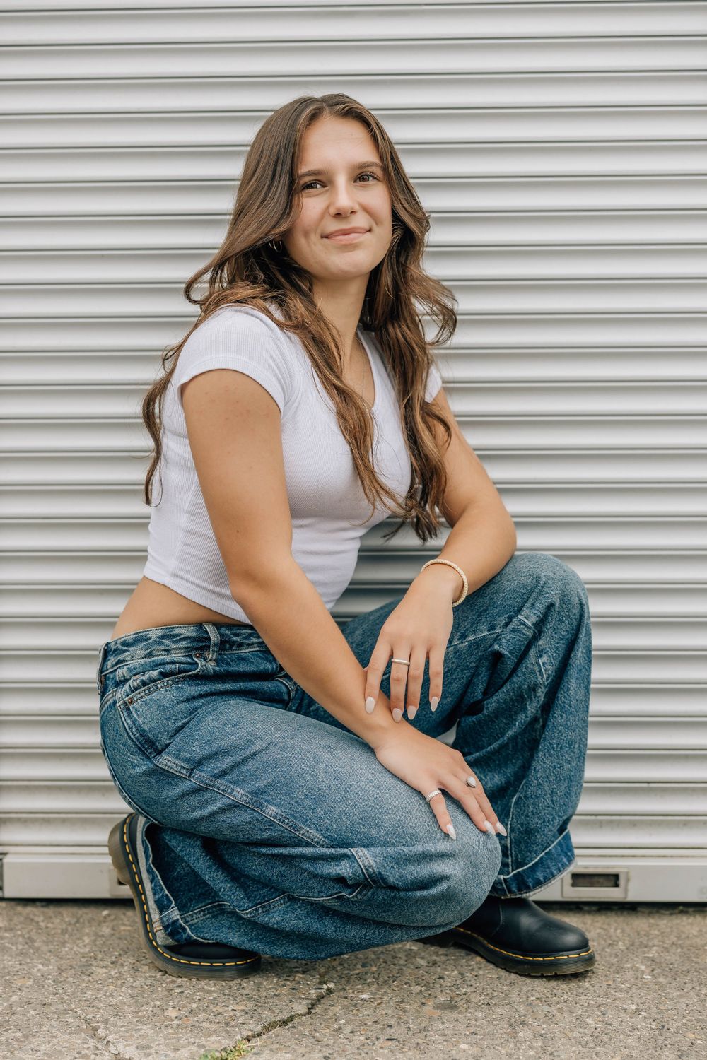 Young woman in white tee and wide-leg jeans crouches in front of a corrugated metal garage door.